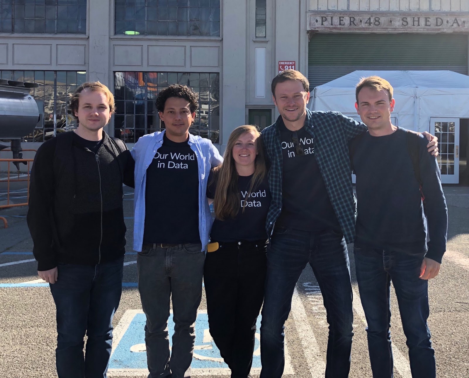 Five people are standing close together, smiling in front of a large building with industrial-style windows. The first person on the left has short, light-colored hair and wears a dark sweater. The second person has curly dark hair, wearing a light blue shirt over a dark shirt. The third person, a woman with long brown hair, is in the center, wearing a dark shirt with the text "Our World in Data." The fourth person is wearing a checkered shirt over a dark shirt and has short hair. The last person on the right has short light hair and is wearing a dark sweater. They are all casually dressed, and the background shows an open space with some structures and a white tent.