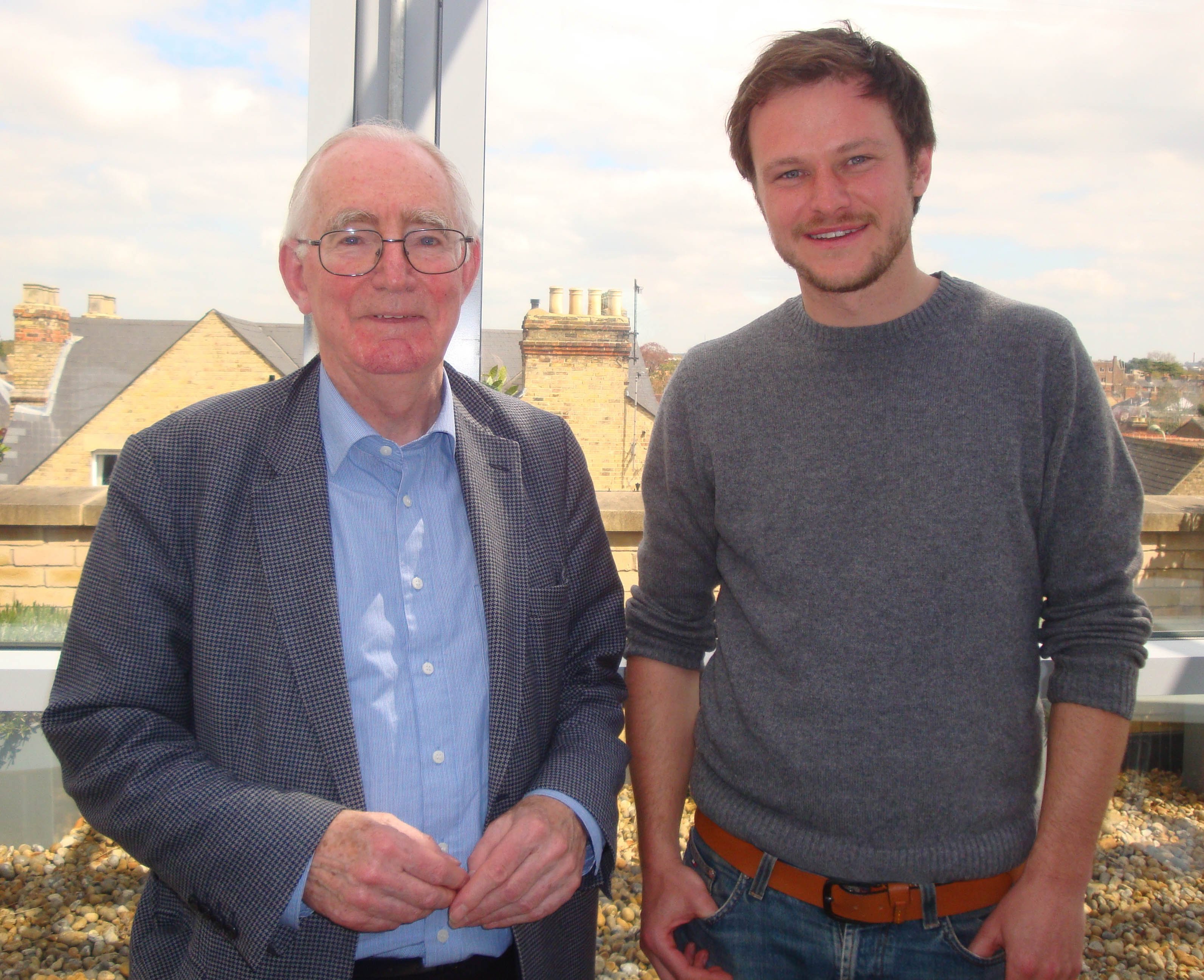 Two men stand together in front of a large window. On the left, an older man with glasses and gray hair wears a checkered blazer over a light blue shirt. He has a gentle smile. On the right, a younger man with short, dark hair and a beard is dressed in a gray sweater and jeans, smiling back at the camera. The background reveals a cityscape with rooftops and a partly cloudy sky. Small pebbles can be seen on the ground in front of them, enhancing the modern and casual atmosphere of the setting.